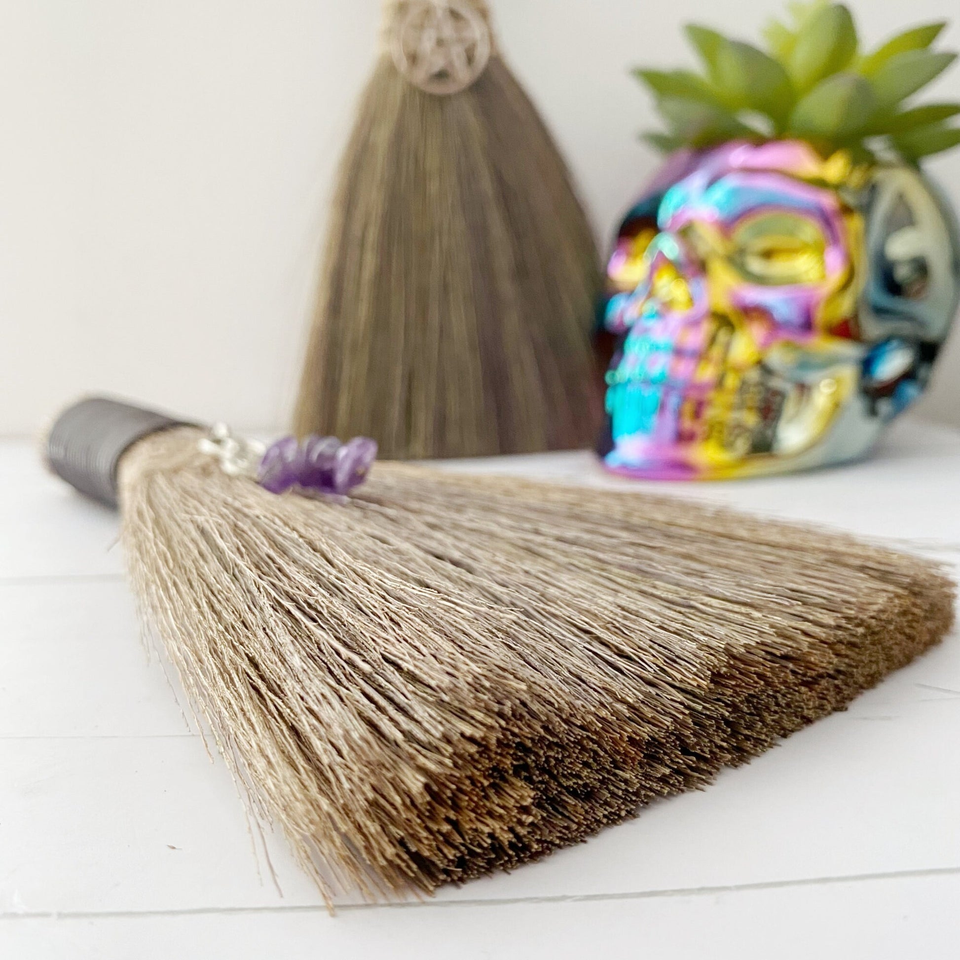 Two Mini Besoms (5 Styles to Select From) with black handles and charms hang against a wall; one charm is an intricate design and the other is a pentagram. Below them are bundled sage sticks and a colorful, reflective skull planter with a green succulent inside.