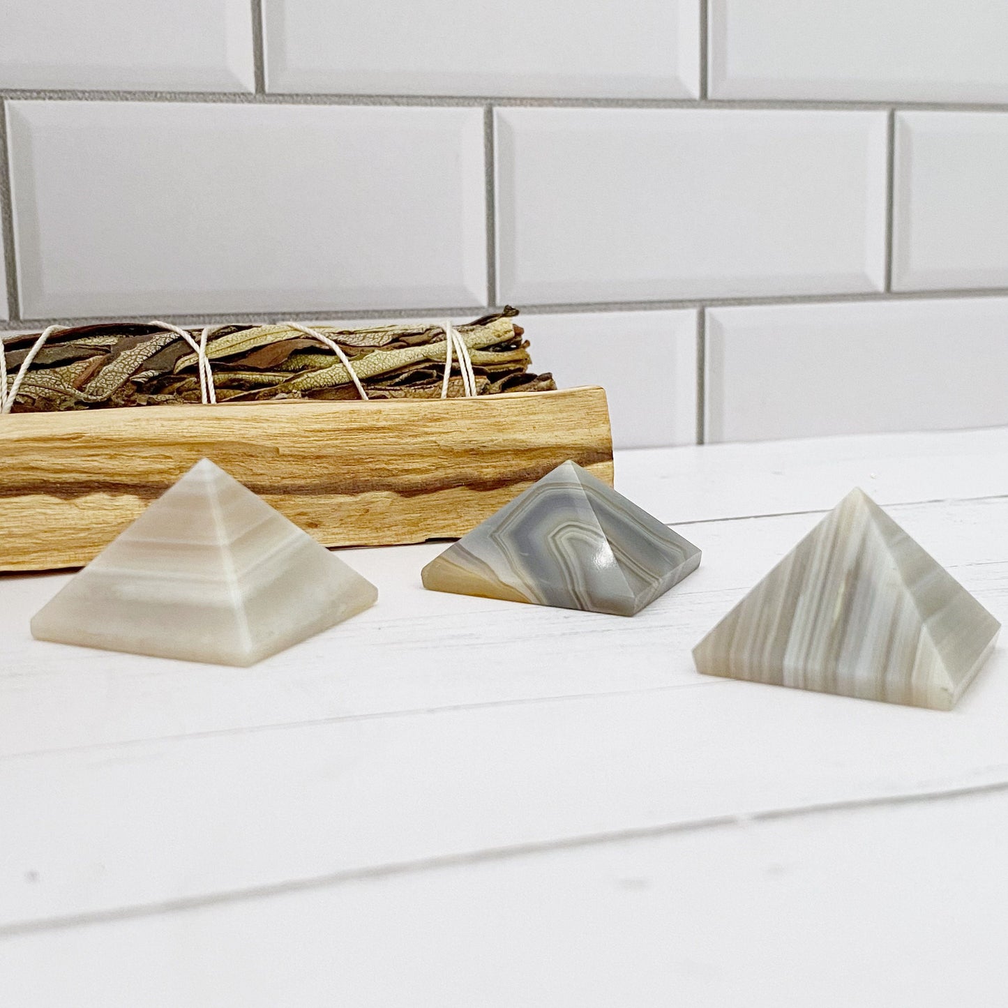 Three small 1" Mini Banded Agate Pyramids, displaying various shades of gray with striped patterns, are placed on a white surface. Behind them is a wooden tray holding a bundle of dried herbs, all set against a white tiled background, creating an atmosphere perfect for rebalancing and harmonizing.