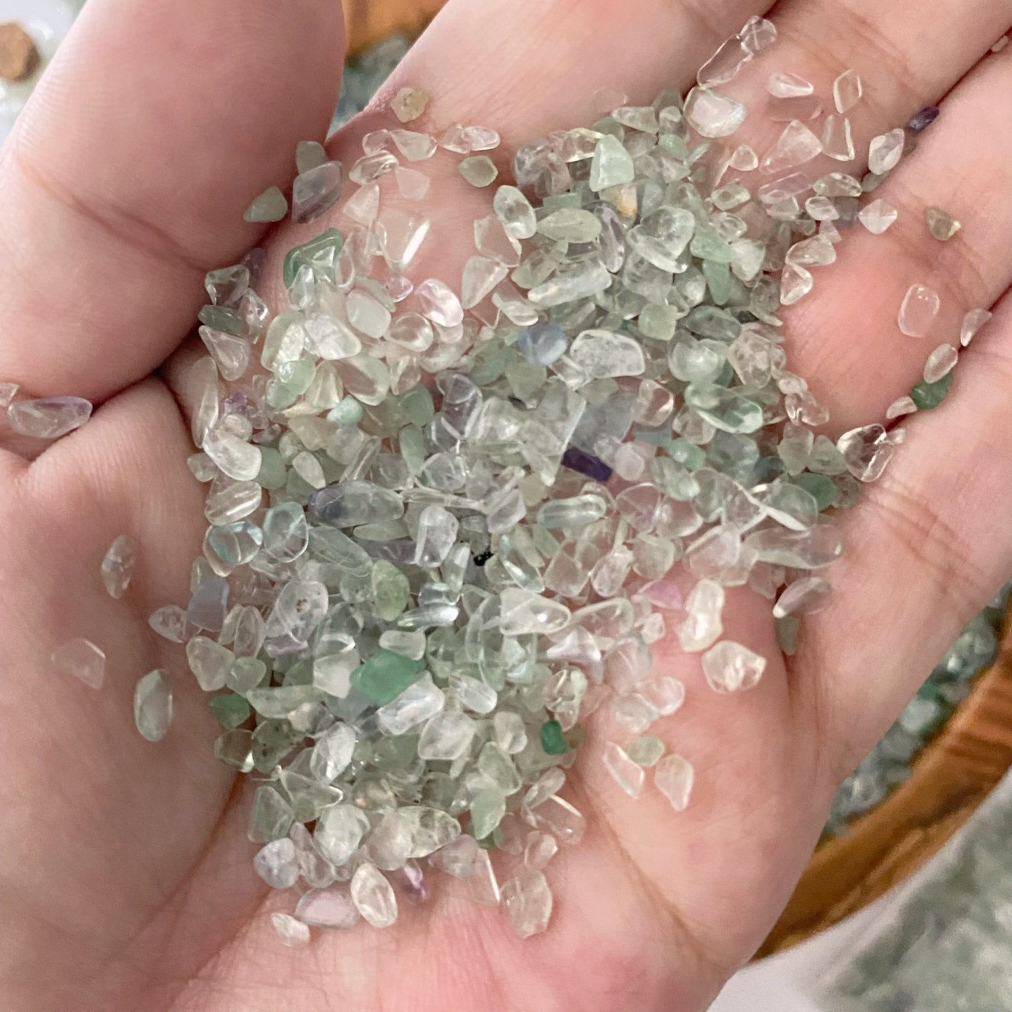 A wooden bowl filled with small, translucent Green Fluorite Crystal Chips is centered on a white surface. Nearby, three small glass bottles also contain Green Fluorite Crystal Chips, and there is a bundle of dried sage in the upper right corner. Some tumbled and polished stones are spilled beside the bowl.