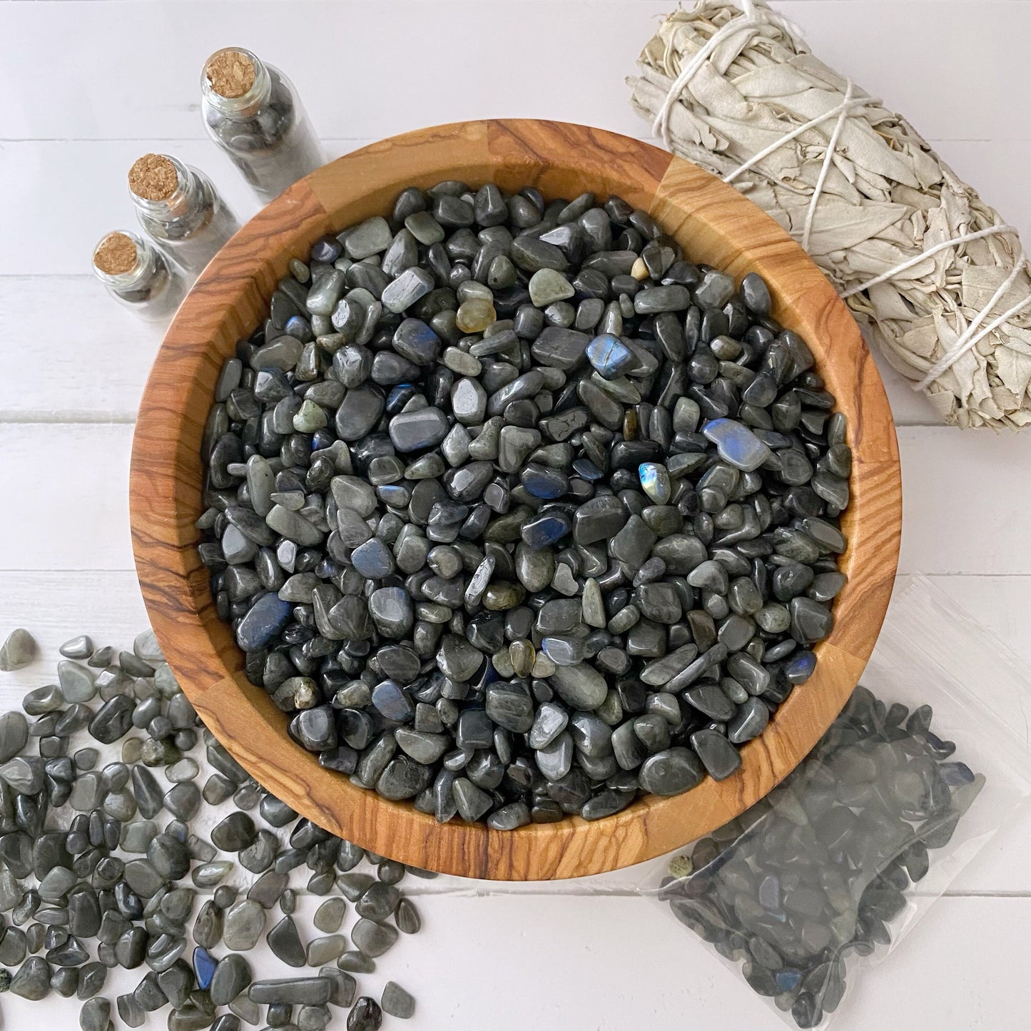 A wooden bowl filled with polished black stones is placed on a white surface. Surrounding the bowl are small glass bottles with cork stoppers, a bundle of white sage, and more black stones scattered around and in a plastic bag. Among the items, Labradorite Crystal Chips shimmer subtly, adding to the mystic allure.