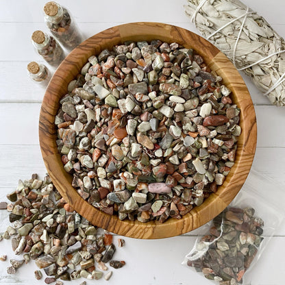 A large wooden bowl filled with polished stones of various colors sits on a white surface. The bowl is surrounded by small jars of Ocean Jasper Crystal Chips, a white sage bundle, and a small plastic bag containing more stones with some scattered on the table, hinting at their metaphysical uses.