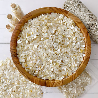 A wooden bowl filled with small, polished Mother of Pearl Crystal Chips sits on a white table. Next to the bowl are bundles of sage wrapped in string and three small glass spell jars containing similar crystals. Some crystals are scattered on the table around the bowl.