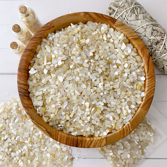 A wooden bowl filled with small, polished Mother of Pearl Crystal Chips sits on a white table. Next to the bowl are bundles of sage wrapped in string and three small glass spell jars containing similar crystals. Some crystals are scattered on the table around the bowl.