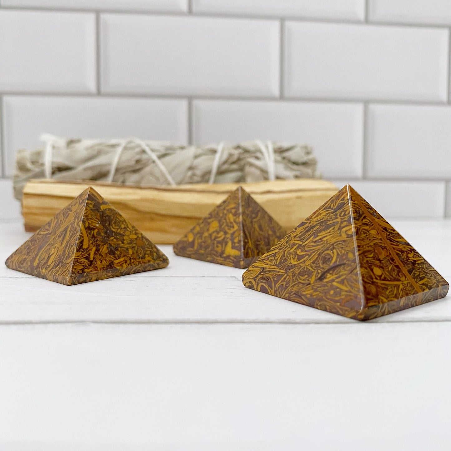 Three small, brown 1" Mini Calligraphy Jasper Pyramids with intricate patterns sit on a white surface in front of a white brick wall. Behind the pyramids, there is a bundle of sage tied with white string, resting on a wooden tray.