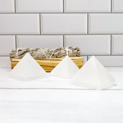 Three polished 1.5" Mini Selenite Pyramids are arranged on a white surface in front of a backdrop of white subway tiles. Behind the pyramids, embodying sacred geometry, is a bundle of dried sage tied with string, resting on a piece of wood. The setting appears calm and minimalistic, perfect for mental clarity.