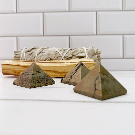 Three 1" Mini Pyrite Pyramids made of a dark stone are placed on a white surface in front of a white tiled background. Behind the pyramids, a bundle of dried sage and a piece of palo santo wood, known for their protective qualities, are set on the surface, aligned horizontally.