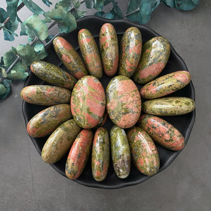 A round black bowl is filled with polished, oval-shaped healing crystals in shades of green and pink. The Unakite Palm Stones are neatly arranged in a radial pattern. Green leaves are visible in the background, adding a natural touch to the display. The surface underneath is grey.