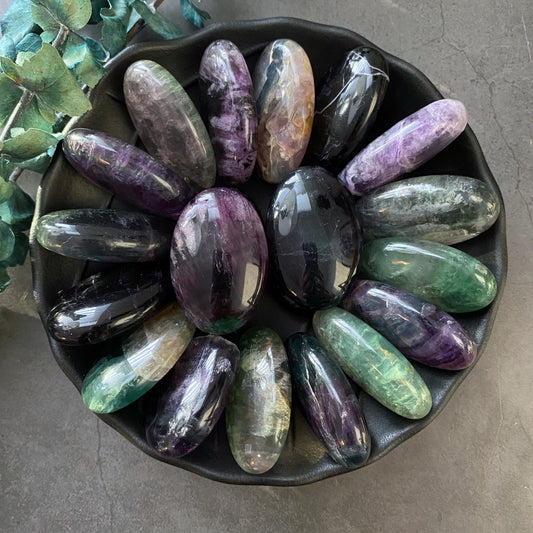 A round, black dish filled with smooth, polished Rainbow Fluorite Palm Stones in various shades of purple, green, and black, arranged in a circular pattern. Emitting harmonizing vibrations and promoting energy and clarity, the stones are accompanied by a few leaves on a dark, textured surface.