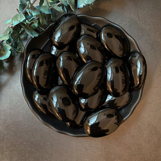 A black bowl filled with glossy Obsidian Palm Stones is placed on a grey surface. Adjacent to the bowl are green eucalyptus leaves. The Obsidian Palm Stones, highly polished, reflect light and highlights from the surrounding environment, offering a sense of mental stress relief.