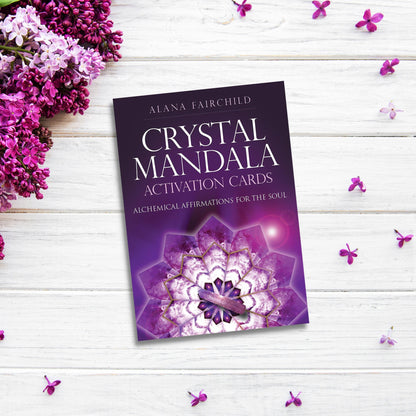 A package of Crystal Mandala Activation Cards resting on a white wooden surface. The intricate purple mandala design is surrounded by purple flowers and scattered petals. Subtext reads "Alchemical Affirmations for the Soul." Ideal for those on a spiritual journey seeking oracle message cards.