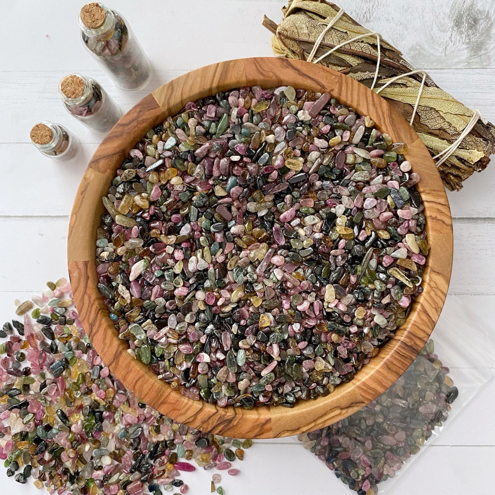 A wooden bowl filled with Rainbow Tourmaline Crystal Chips is surrounded by small glass jars containing similar stones, a bundle of sage tied with string, and additional gemstones scattered on a white surface. Perfect for those seeking metaphysical crafting supplies.