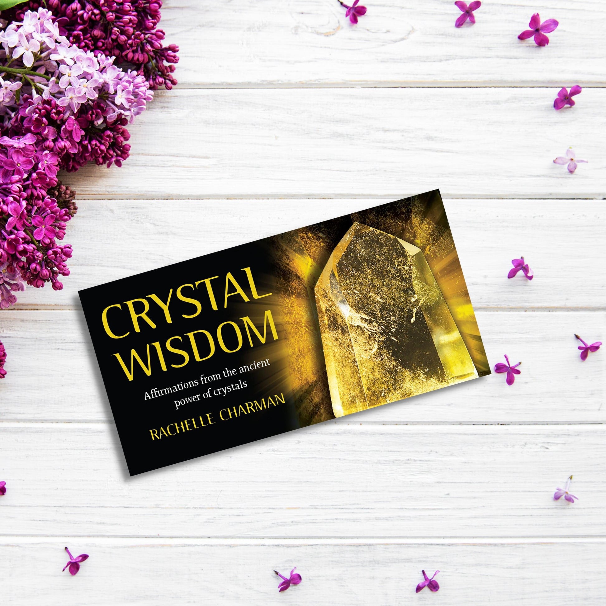 A product titled "Crystal Wisdom Cards" by Rachelle Charman is placed on a white wooden surface with clusters of purple flowers and scattered small purple petals. The product cover, reminiscent of soothing self-healing meditation, features an image of a crystal with a golden glow.