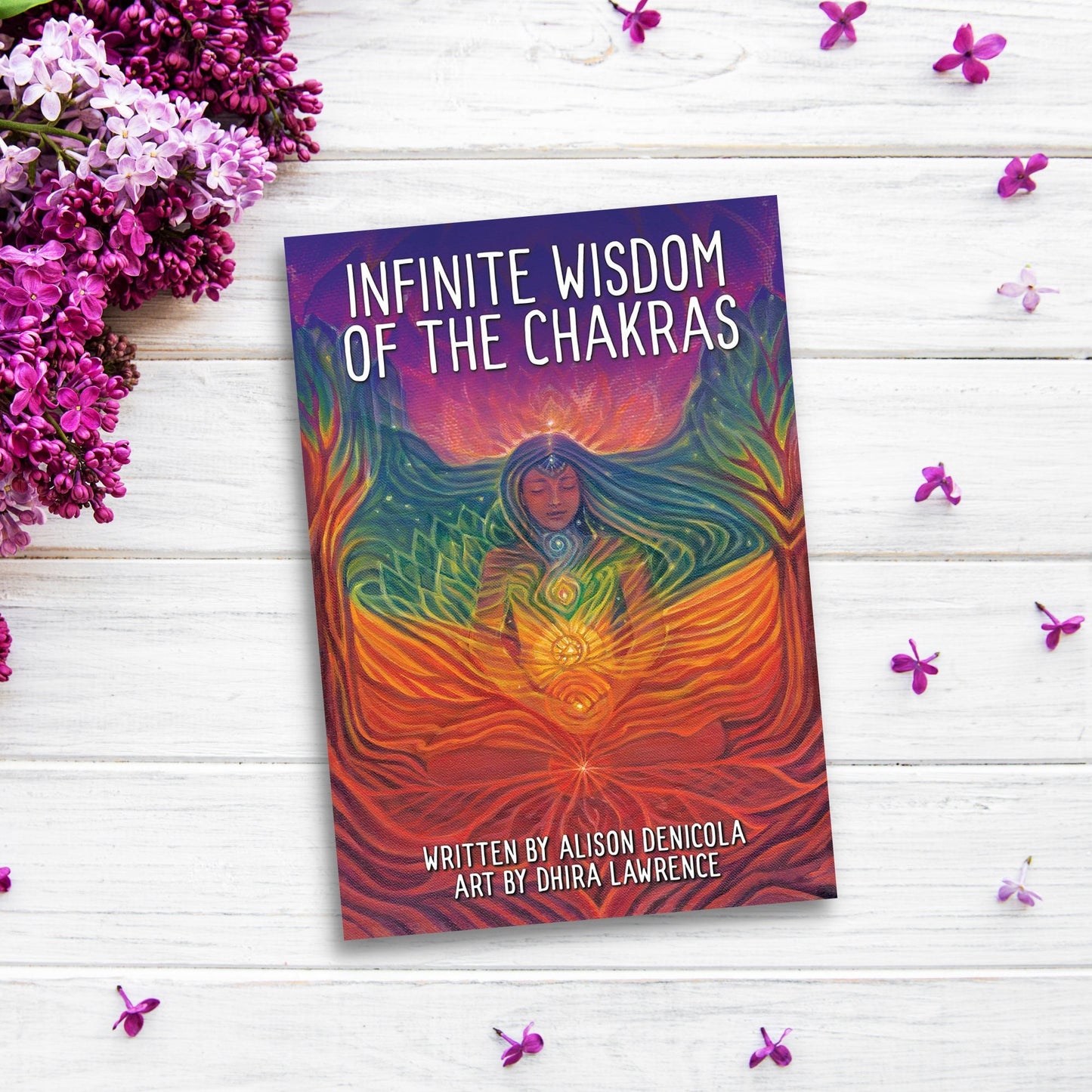 Infinite Wisdom of the Chakras, written by Alison Denicola with art by Dhira Lawrence, is placed on a white wooden surface surrounded by purple flowers. The cover features a meditative figure with vibrant, colorful chakra energy, beautifully illustrating the healing power of the chakra system.