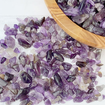 A wooden bowl filled with small polished Amethyst Crystal Chips is placed on a white surface. Surrounding the bowl are scattered amethyst stones, small bottles containing crystals, and a bundle of white sage to enhance psychic abilities. A plastic bag of stones is also visible.