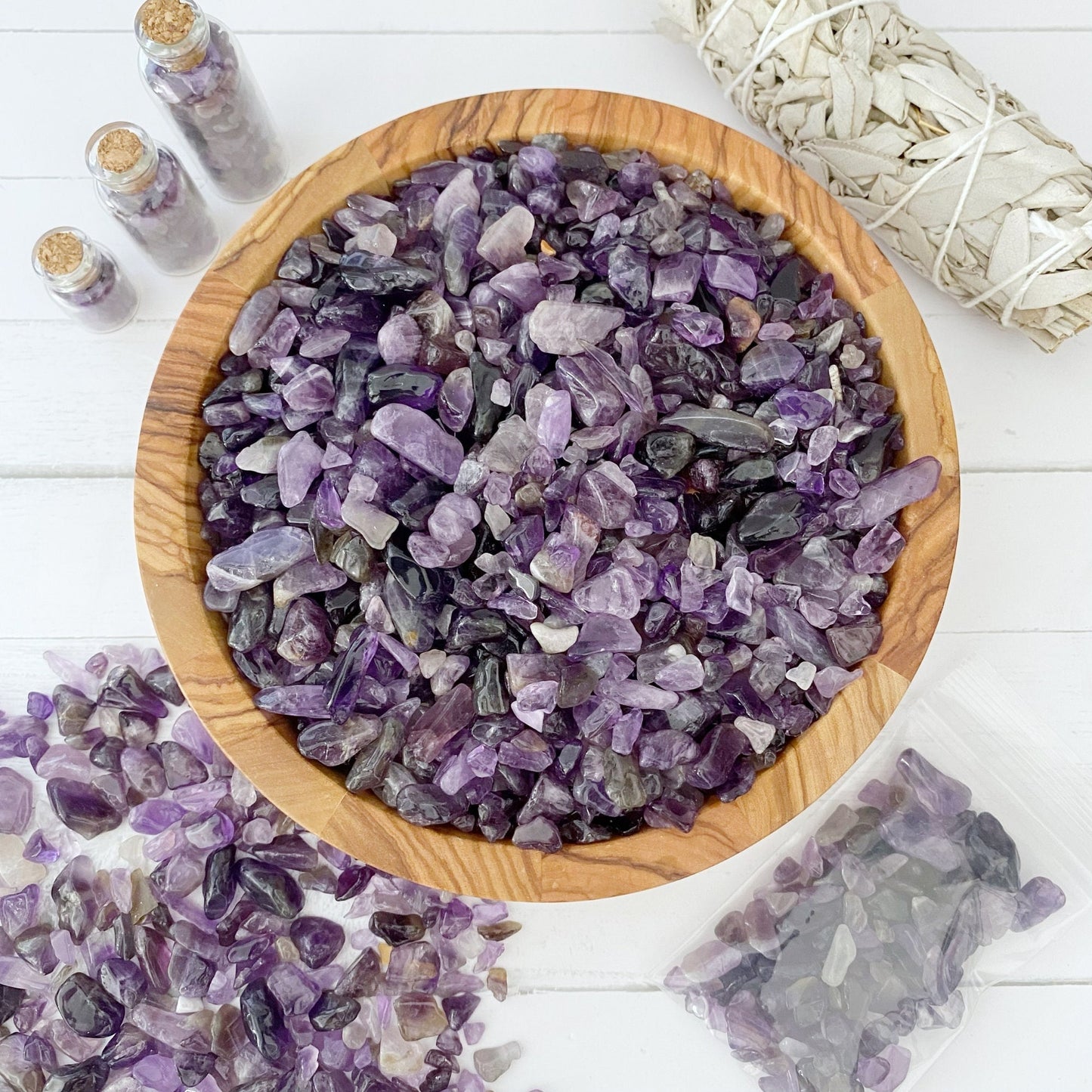 A wooden bowl filled with small polished Amethyst Crystal Chips is placed on a white surface. Surrounding the bowl are scattered amethyst stones, small bottles containing crystals, and a bundle of white sage to enhance psychic abilities. A plastic bag of stones is also visible.