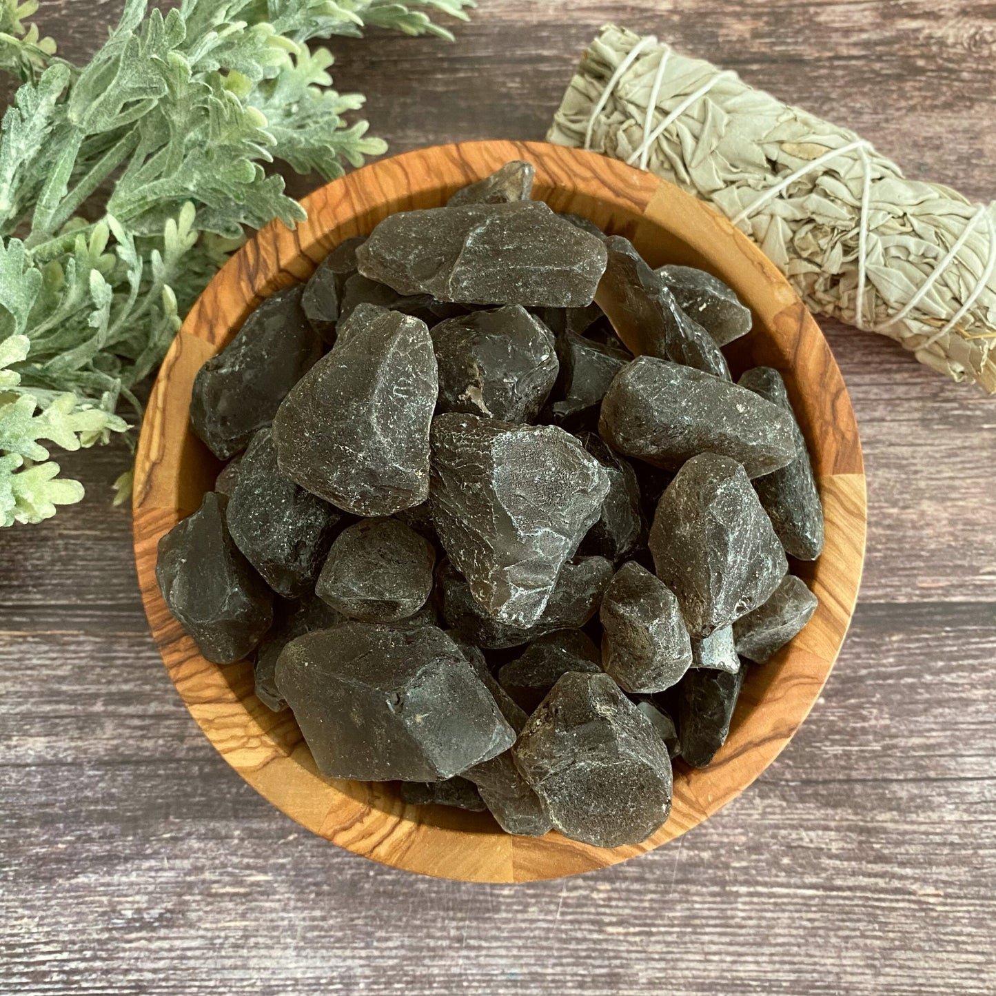 A wooden bowl filled with Rough Raw Smoky Quartz Stones is placed on a wooden surface. To the right, there is a bundle of dried sage wrapped in twine. On the left, some green leafy plant foliage is visible. The overall composition has a rustic and natural feel, ideal for manifestation practices.