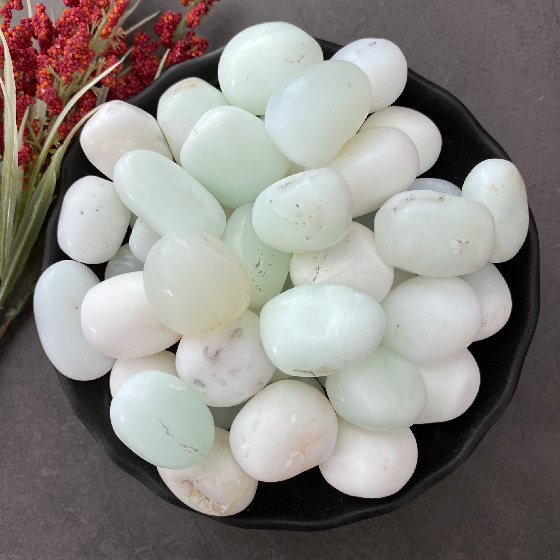 A black bowl filled with smooth, polished, semi-translucent White Jade Tumbled Stones and light green stones. Red and yellow dried flowers are placed to the side for decoration, set on a dark gray background. The serene arrangement promotes internal harmony and dispels negative thinking.