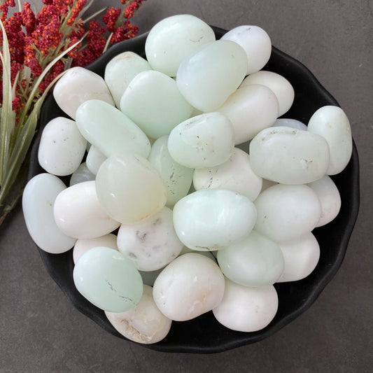 A black bowl filled with smooth, polished, semi-translucent White Jade Tumbled Stones and light green stones. Red and yellow dried flowers are placed to the side for decoration, set on a dark gray background. The serene arrangement promotes internal harmony and dispels negative thinking.