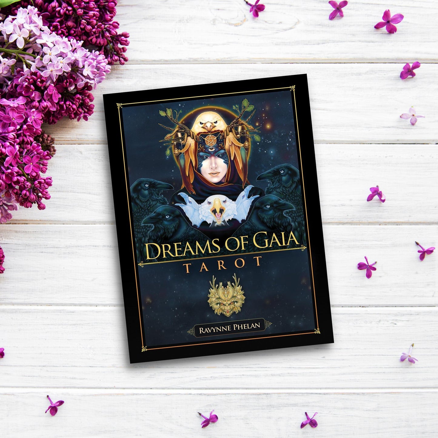 The "Dreams of Gaia Tarot" lies on a white wooden surface with small purple flowers scattered around. The cover, part of this enchanting tarot card deck, features mystical artwork of a woman with a headdress, two black birds, and a glowing central symbol.