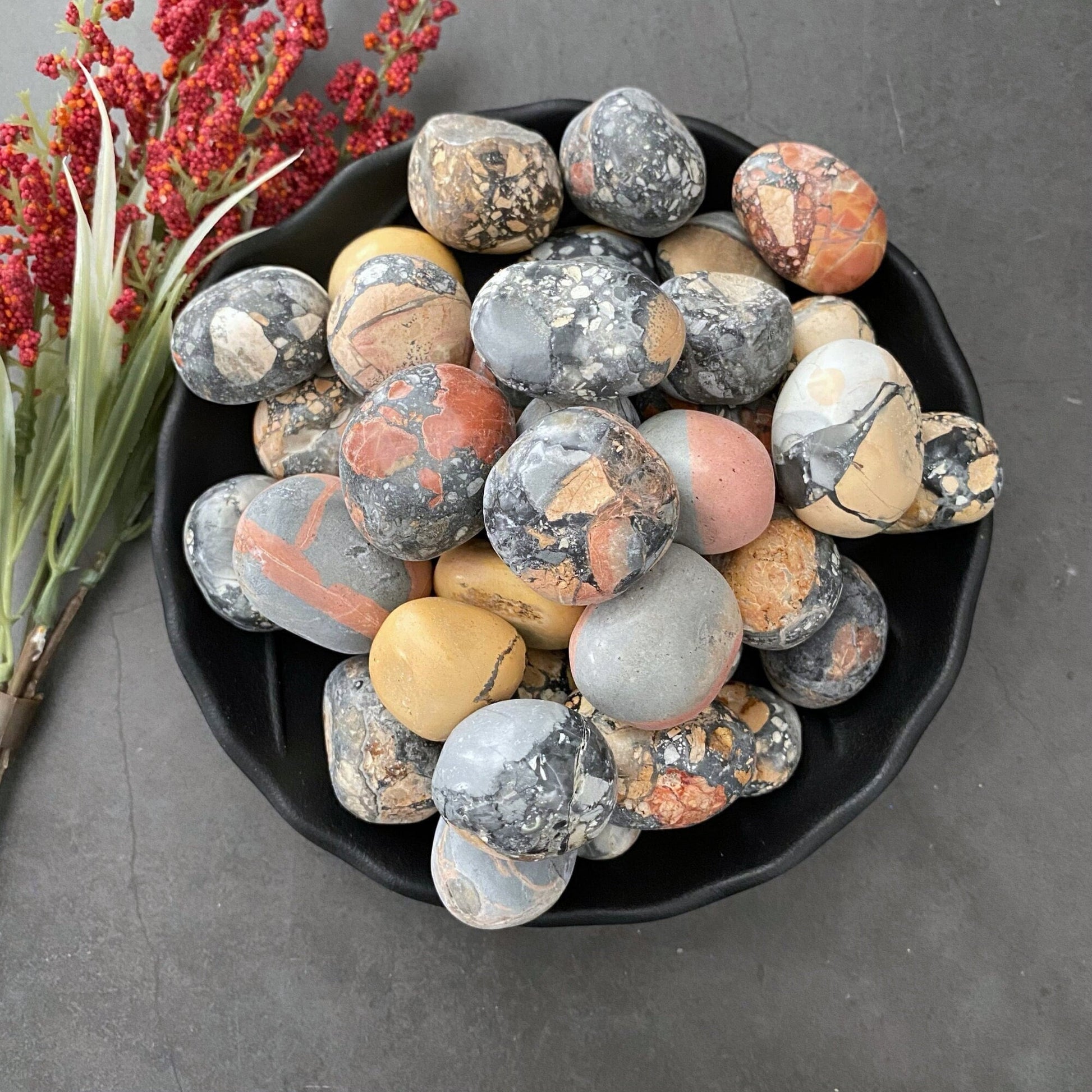 A black bowl filled with hand-polished, multicolored stones sits on a dark, textured surface. The stones have various patterns and shades, including gray, orange, yellow, and pink. Red and green foliage lies next to the bowl, highlighting a stunning Maligano Jasper Tumbled Stones among the mix.