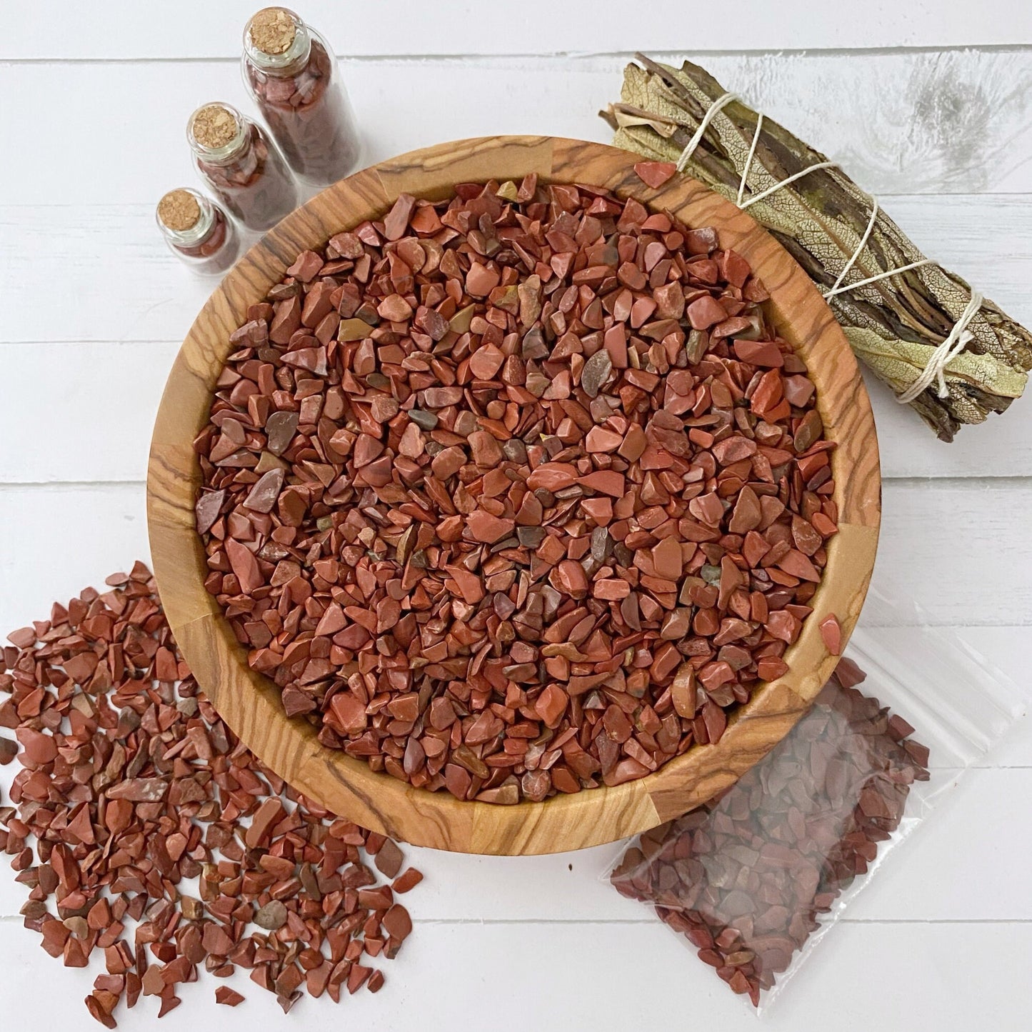 A wooden bowl filled with small red stones is placed on a white surface. Three small bottles filled with Red Jasper Crystal Chips, a bundle of leaves tied with string, and a small plastic bag with more stones are arranged around the bowl, perfect for those who cherish crafting supplies and metaphysical properties.