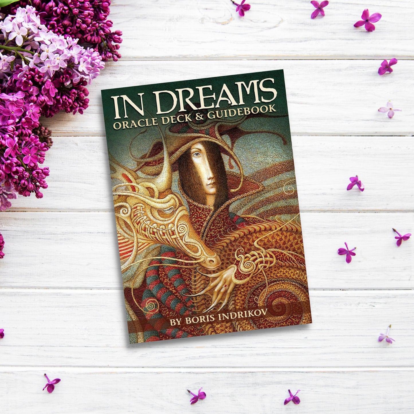 A book titled "In Dreams Oracle Deck" by Boris Indrikov is displayed on a white wooden surface surrounded by purple flowers. The cover features a mystical illustration of a figure blending with intricate patterns and a dragon, perfect for those interested in dream divination.