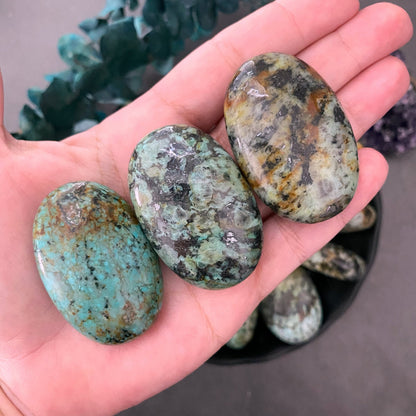 A circular arrangement of polished green and black African Turquoise Palm Stones surrounding a single larger stone in the center, displayed on a dark plate. The background also features a purple amethyst cluster and sprigs of green leaves. The surface is a gray stone texture, enhancing the self-development theme.