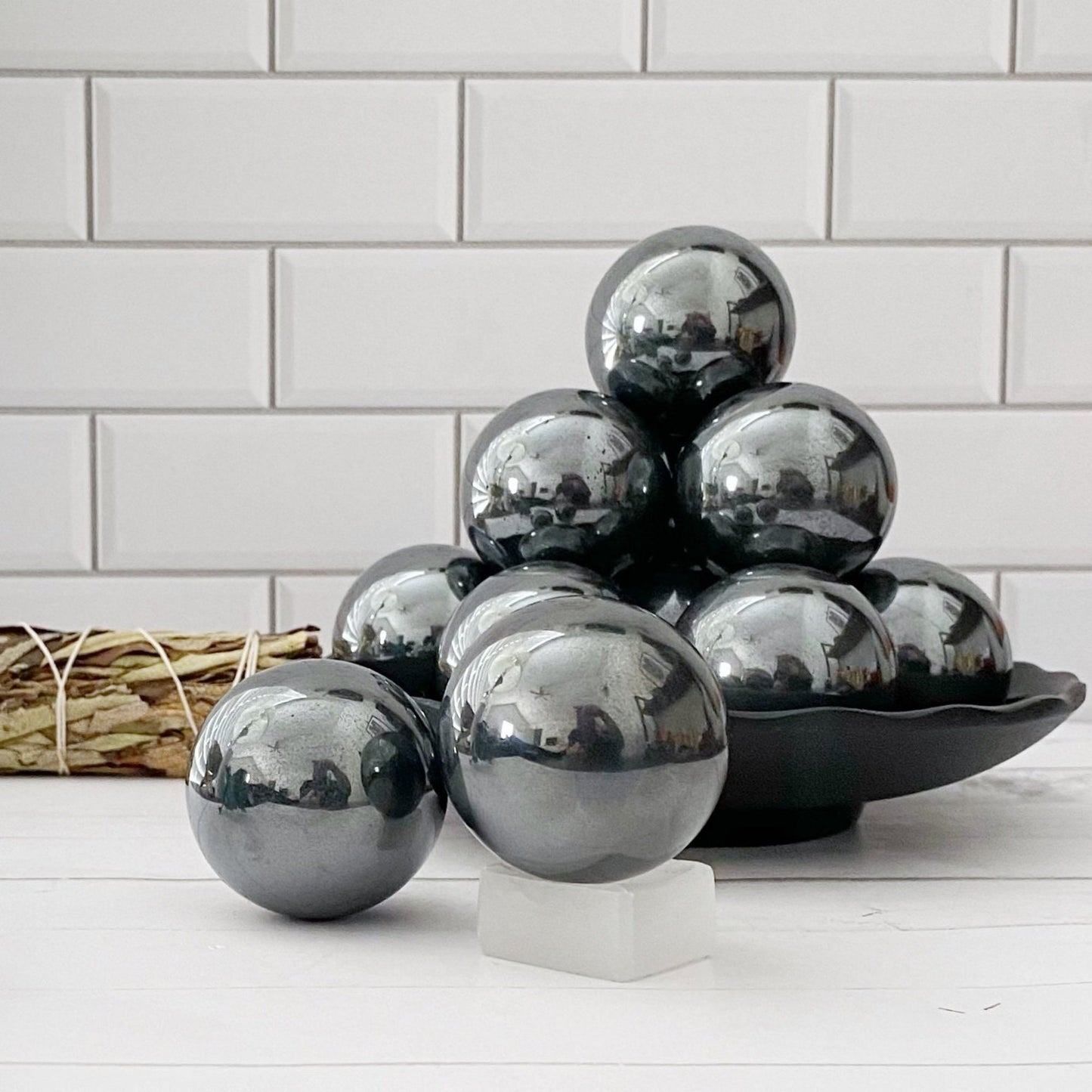 A cluster of reflective metallic balls, likely 40mm Hematite Spheres with their grounding properties, is displayed against a white subway tile background. The spheres are arranged on a dish, capturing reflections of the room and a person on their polished surfaces. A small white cube sits in the foreground.