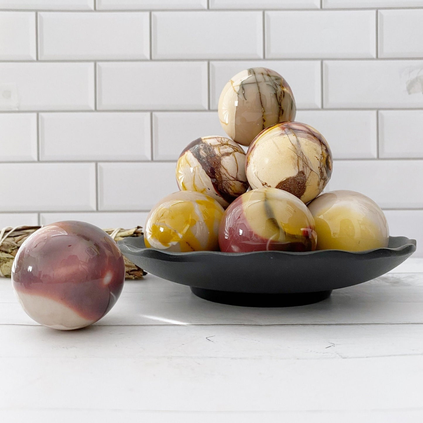 A black plate holds a stack of marbled eggs reminiscent of a 40mm Mookaite Crystal Ball, showcasing a blend of white, yellow, brown, and maroon shades. One marbled egg sits separately on a white countertop. The background includes white subway tiles.