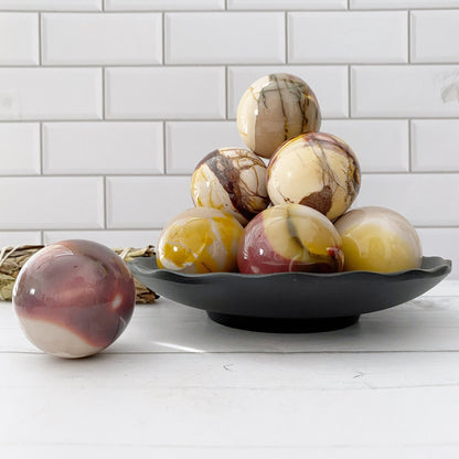 A black plate holds a stack of marbled eggs reminiscent of a 40mm Mookaite Crystal Ball, showcasing a blend of white, yellow, brown, and maroon shades. One marbled egg sits separately on a white countertop. The background includes white subway tiles.