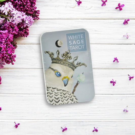 A pocket-sized White Sage Tarot in Tin lies on a white wooden surface, surrounded by small purple flowers and a bouquet of lilacs. The tarot deck features a crowned white bird with a scepter on the cover, accented with chakra colors.