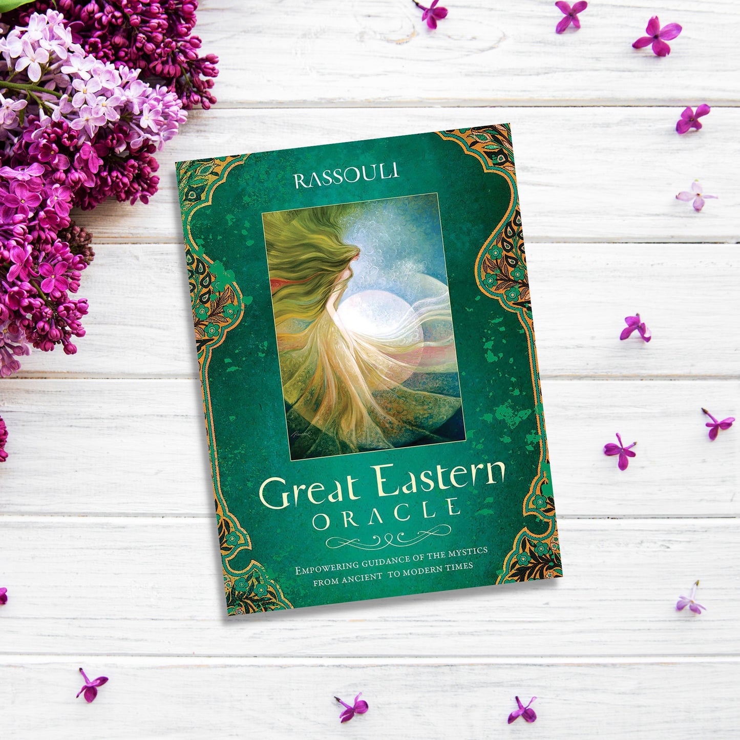 The image shows the "Great Eastern Oracle" placed on a white wooden surface. The deck's box features a rich green and gold design with mystical imagery, perfect for divination. Scattered purple flowers and petals add vibrant contrast, enhancing the scene’s spiritual enrichment.