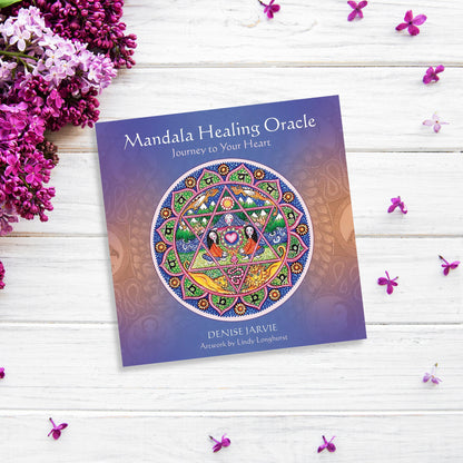 A book titled "Mandala Healing Oracle" by Denise Jarvie with artwork by Lindy Longhurst on a light wooden surface, surrounded by purple and pink flowers. The cover features an intricate, colorful mandala design, offering healing and inspiration through its guidebook with meditations.