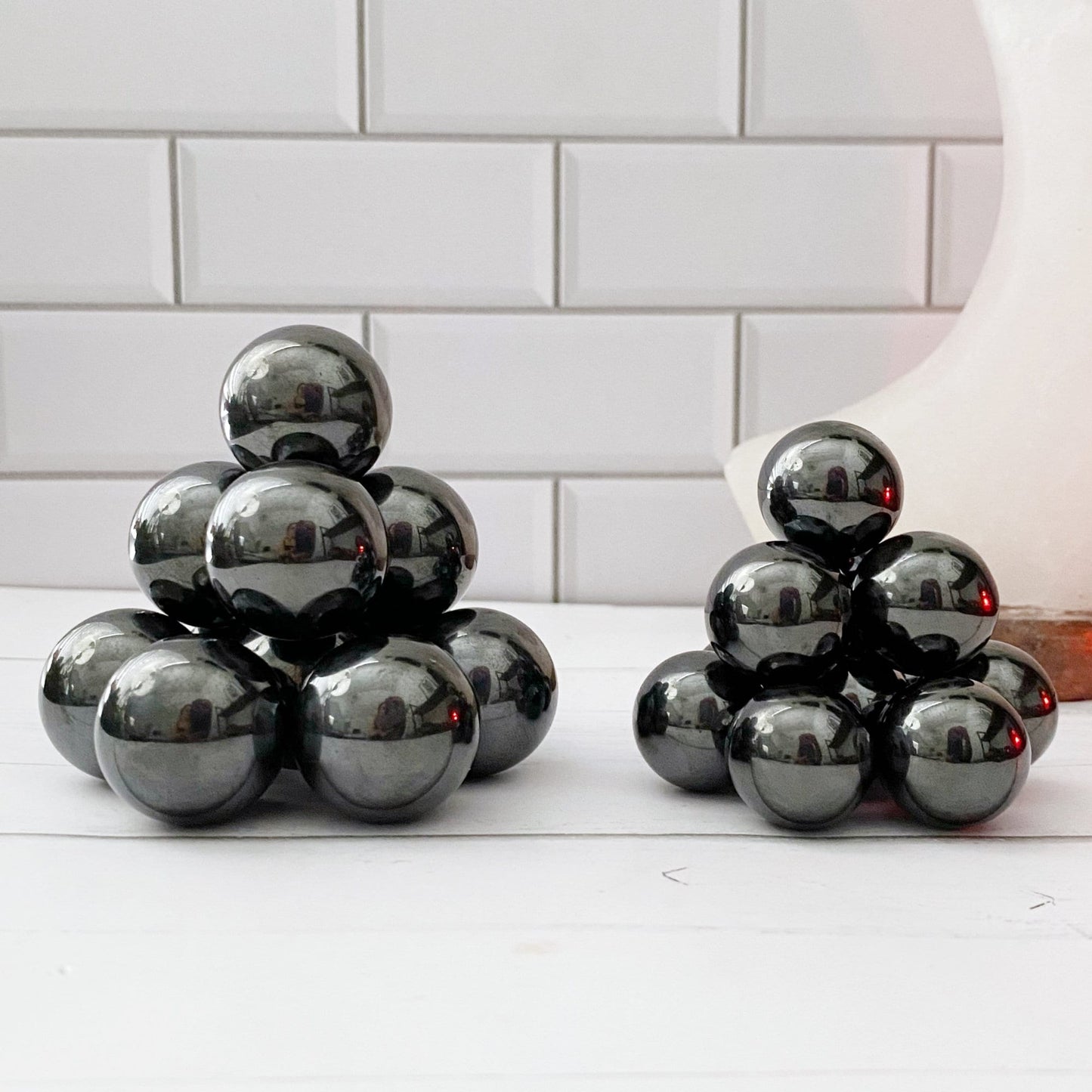 Two pyramid-like structures made from shiny Mini Hematite Zingers are arranged on a white surface. The background features white, rectangular tiles with thin grout lines. A partially visible, smooth, white and beige object is on the right side of the image, suggesting potential meditation tools or stress relief items nearby.