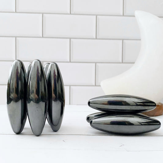 A set of shiny, oval-shaped magnetic toys, known as Hematite Rattlesnake Eggs - Elliptical Olive Zingers, are displayed on a white surface. They are arranged into two groupings: five stacked vertically in a ring formation on the left and two stacked horizontally on the right. A tiled white wall is in the background.