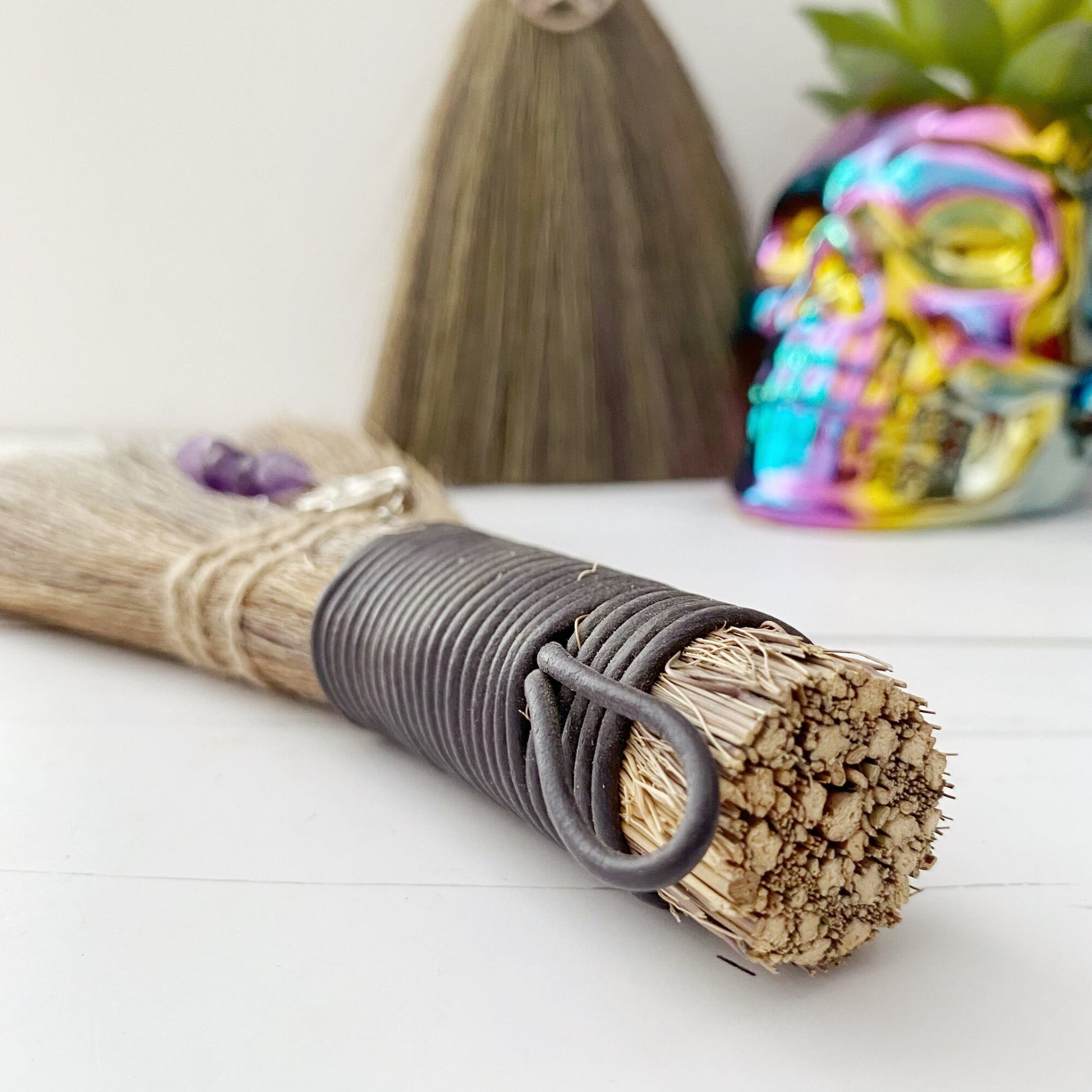 Two Mini Besoms (5 Styles to Select From) with black handles and charms hang against a wall; one charm is an intricate design and the other is a pentagram. Below them are bundled sage sticks and a colorful, reflective skull planter with a green succulent inside.