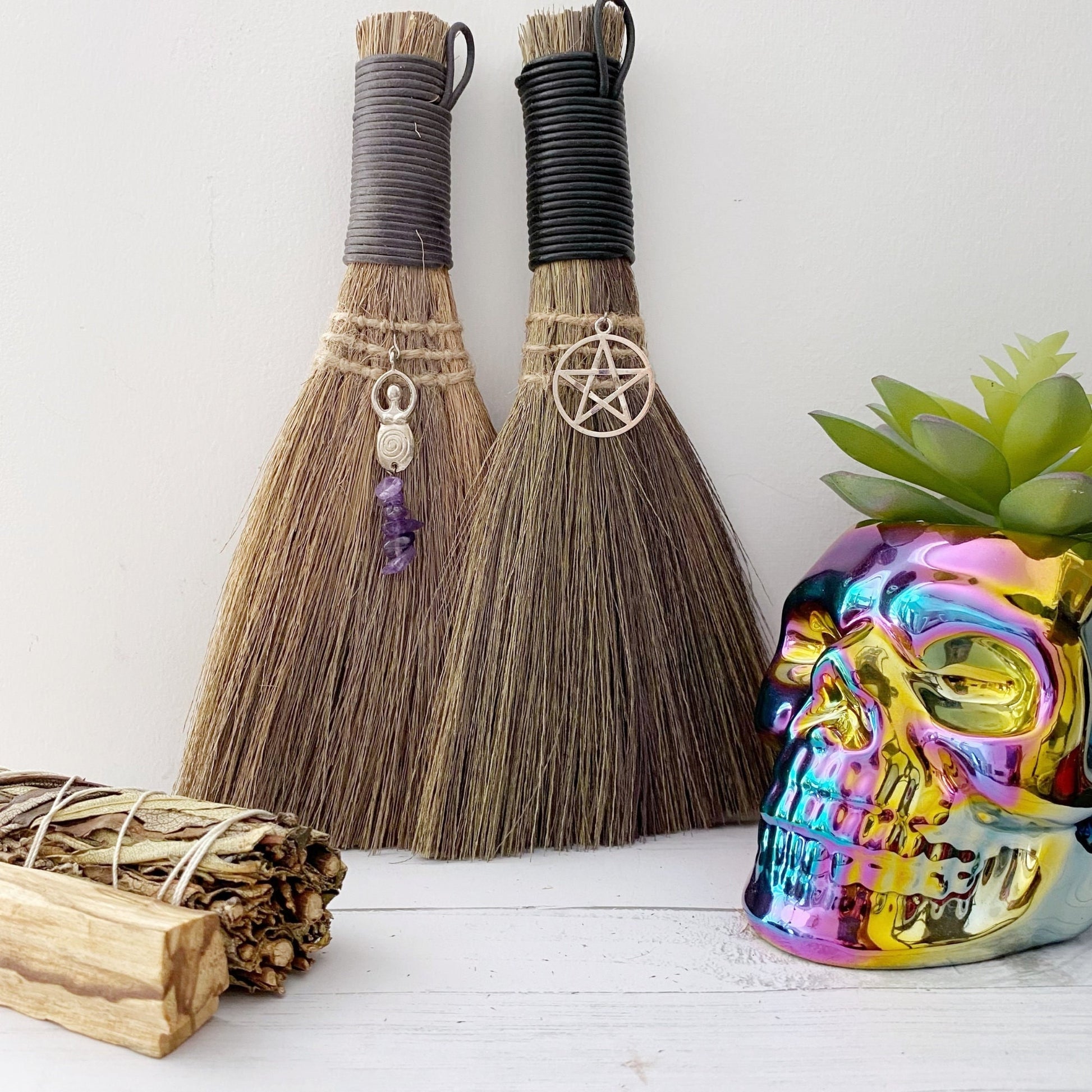 Two Mini Besoms (5 Styles to Select From) with black handles and charms hang against a wall; one charm is an intricate design and the other is a pentagram. Below them are bundled sage sticks and a colorful, reflective skull planter with a green succulent inside.