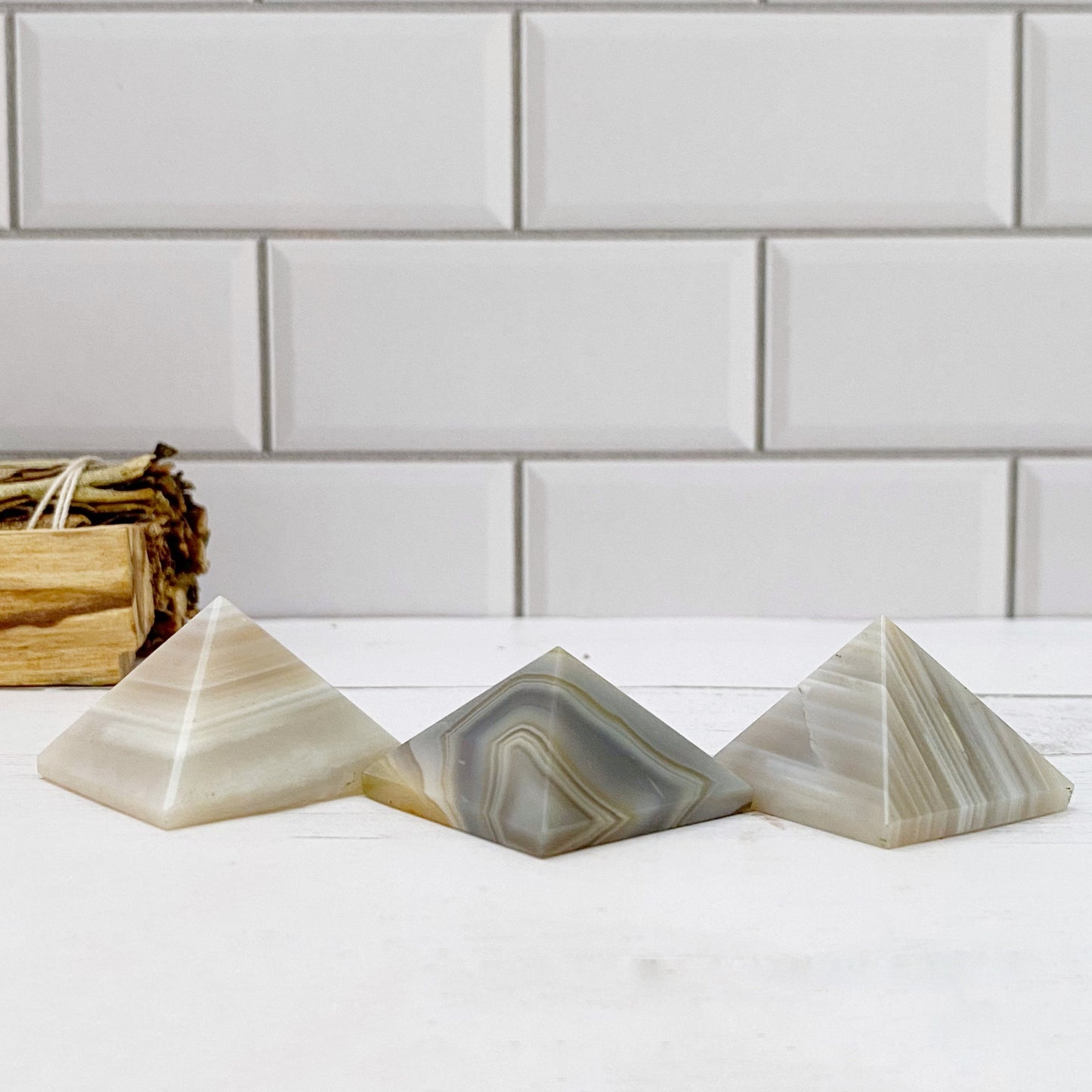 Three small 1" Mini Banded Agate Pyramids, displaying various shades of gray with striped patterns, are placed on a white surface. Behind them is a wooden tray holding a bundle of dried herbs, all set against a white tiled background, creating an atmosphere perfect for rebalancing and harmonizing.