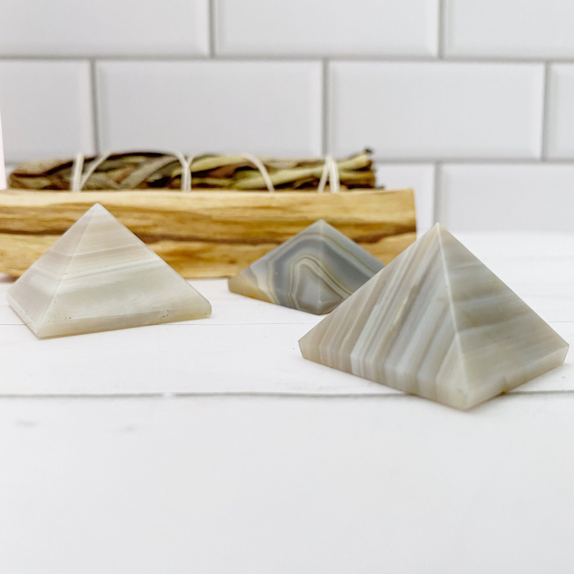 Three small 1" Mini Banded Agate Pyramids, displaying various shades of gray with striped patterns, are placed on a white surface. Behind them is a wooden tray holding a bundle of dried herbs, all set against a white tiled background, creating an atmosphere perfect for rebalancing and harmonizing.