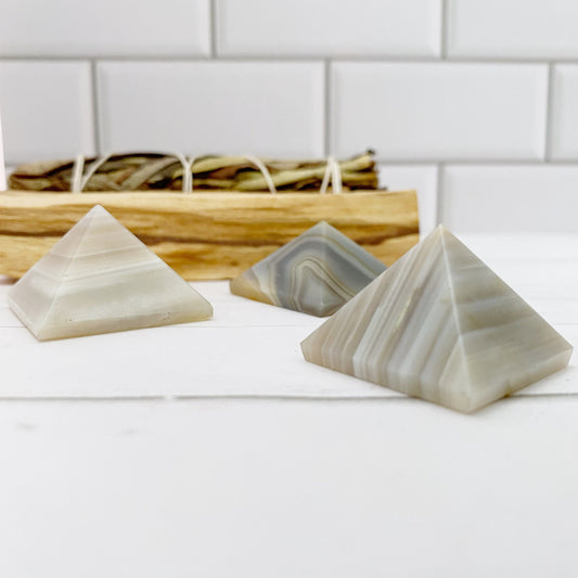 Three small 1" Mini Banded Agate Pyramids, displaying various shades of gray with striped patterns, are placed on a white surface. Behind them is a wooden tray holding a bundle of dried herbs, all set against a white tiled background, creating an atmosphere perfect for rebalancing and harmonizing.