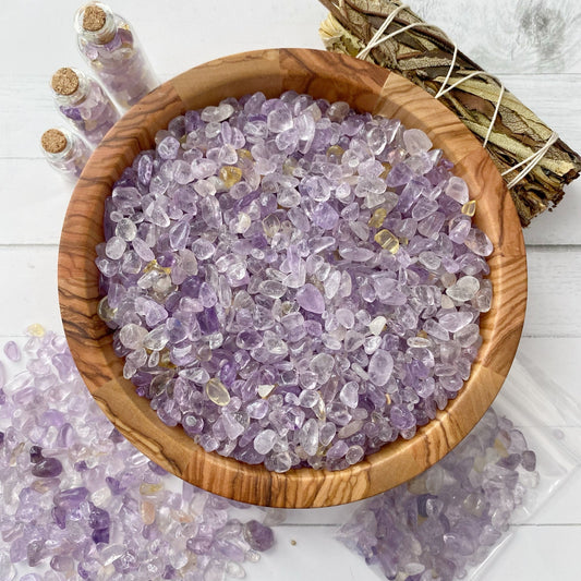 A wooden bowl filled with small, polished pieces of amethyst gemstones is placed on a white surface. Three small glass vials containing similar stones and colorful Ametrine Crystal Chips are in the background, along with a sage smudge stick and a scattered pile of stones.