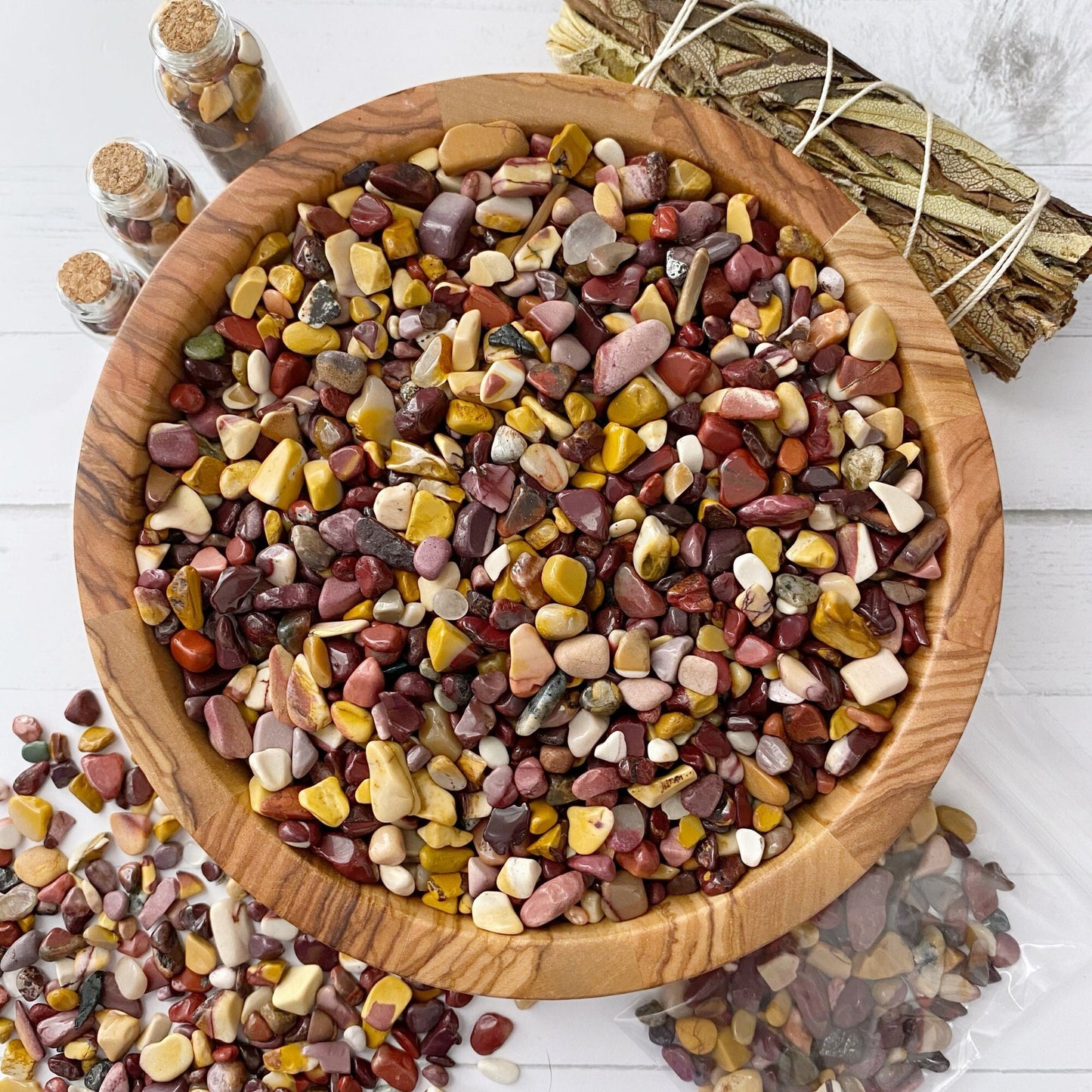 A wooden bowl filled with tumbled and polished Mookaite Crystal Chips is placed on a white surface. Nearby, small glass jars filled with Mookaite Crystal Chips and a bundle of sage sticks are also visible. Some of the stones have spilled out of the bowl and are scattered around it, perfect for your next metaphysical crafting session.