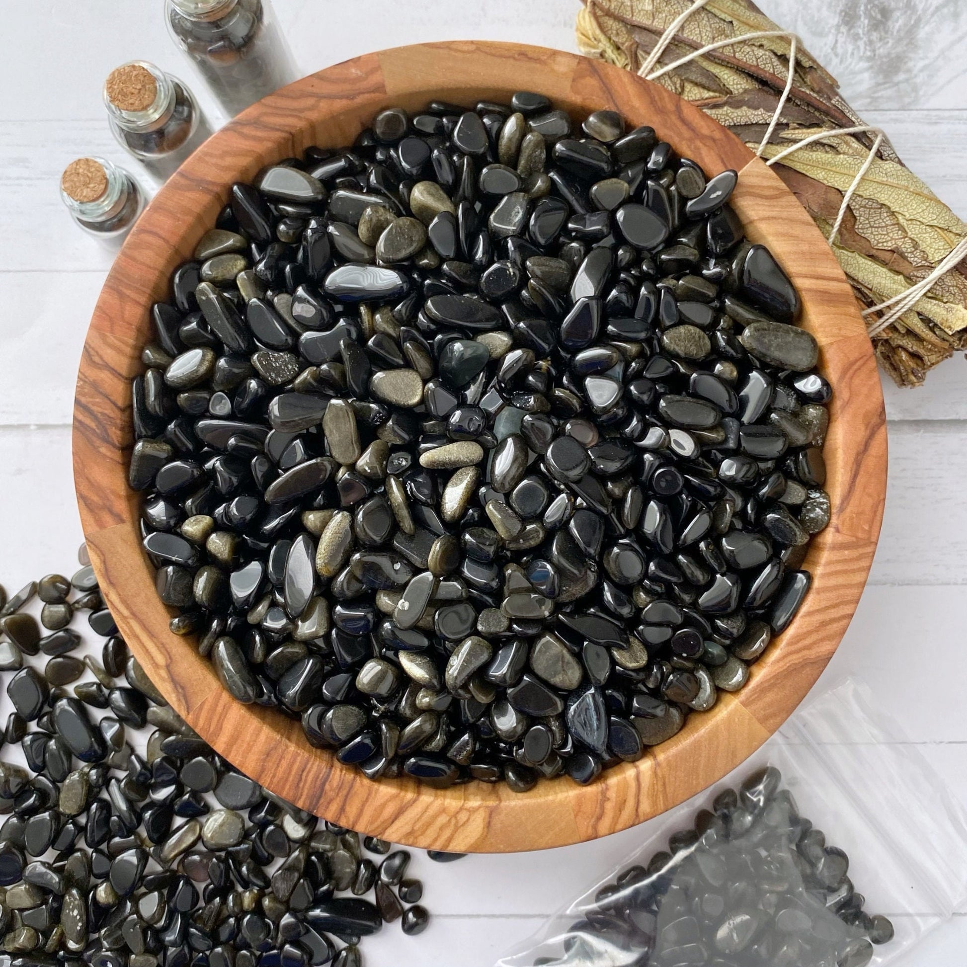 A wooden bowl filled with shiny, polished Gold Sheen Obsidian Crystal Chips is surrounded by similar stones spilling out onto a surface. To the side, there are corked glass bottles, a wrapped bundle of dried leaves, and a clear plastic bag containing more of these psychic protection stones.