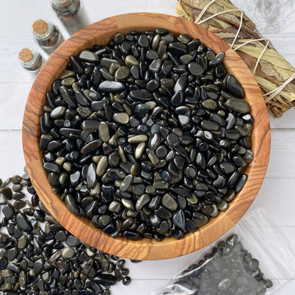 A wooden bowl filled with shiny, polished Gold Sheen Obsidian Crystal Chips is surrounded by similar stones spilling out onto a surface. To the side, there are corked glass bottles, a wrapped bundle of dried leaves, and a clear plastic bag containing more of these psychic protection stones.