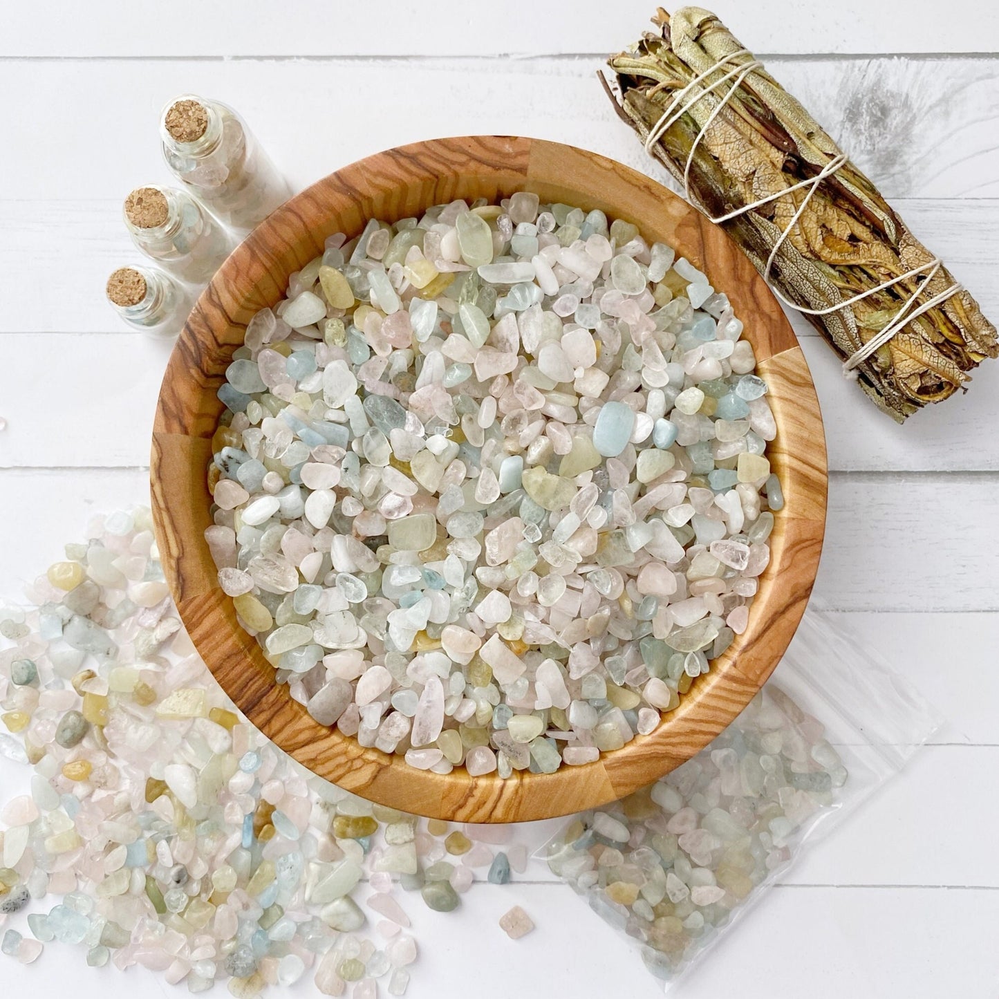 A wooden bowl filled with an assortment of small, tumbled polished stones sits on a white surface. Beside the bowl are three small corked glass bottles, a bundle of sage, and a plastic bag containing Morganite Crystal Chips. Some loose stones are scattered around the bowl.