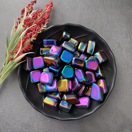 A black bowl filled with shiny, magical Rainbow Hematite Tumbled Stones that display various metallic colors such as purple, blue, green, and gold. Next to the bowl is a small bouquet of red berries and green leaves placed on a dark surface.