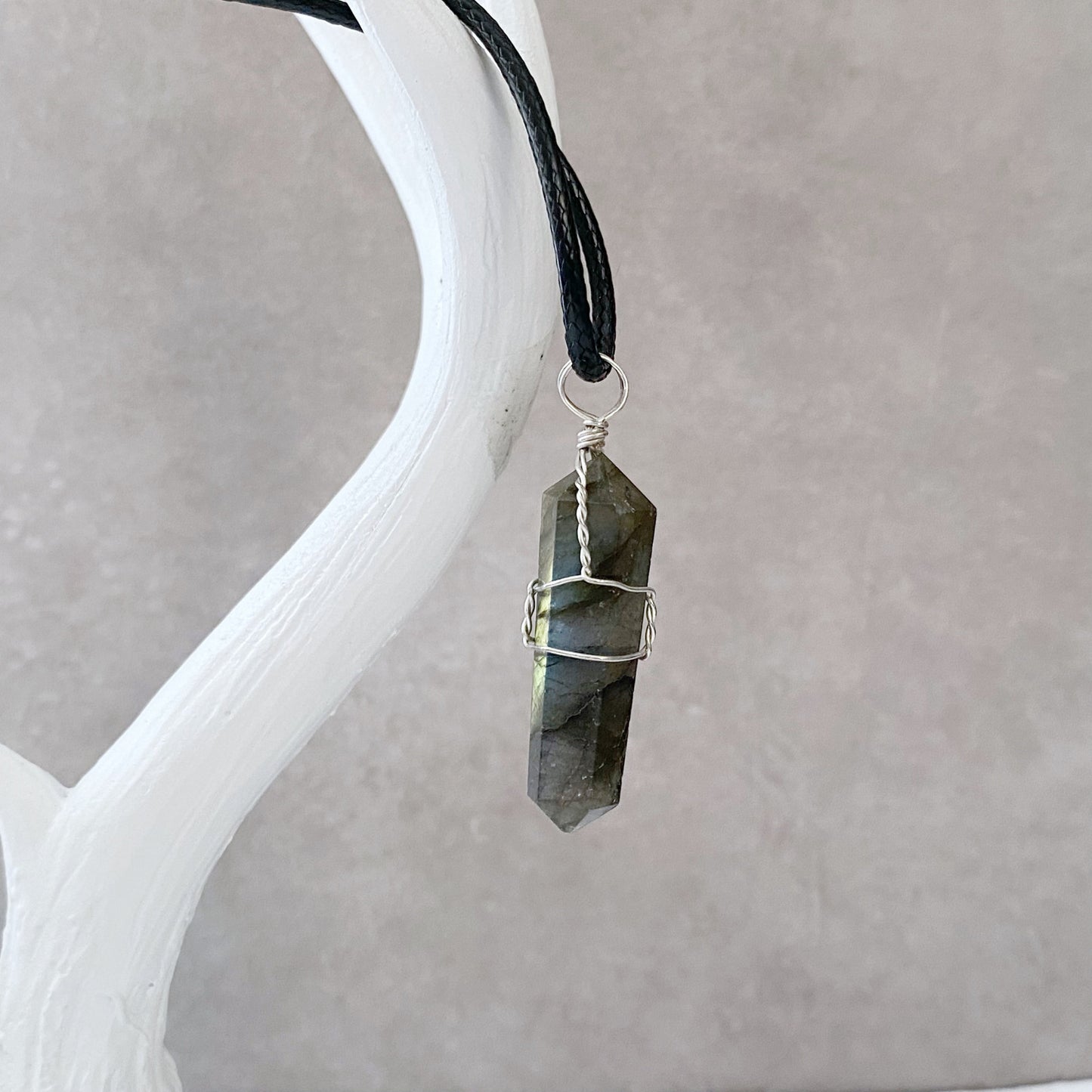 A 1.5" Double Terminated Labradorite Point Pendant wrapped in silver wire, attached to a black cord, is displayed against a soft gray background. The crystal has iridescent shades of blue and green and hangs elegantly from a white antler-shaped holder.