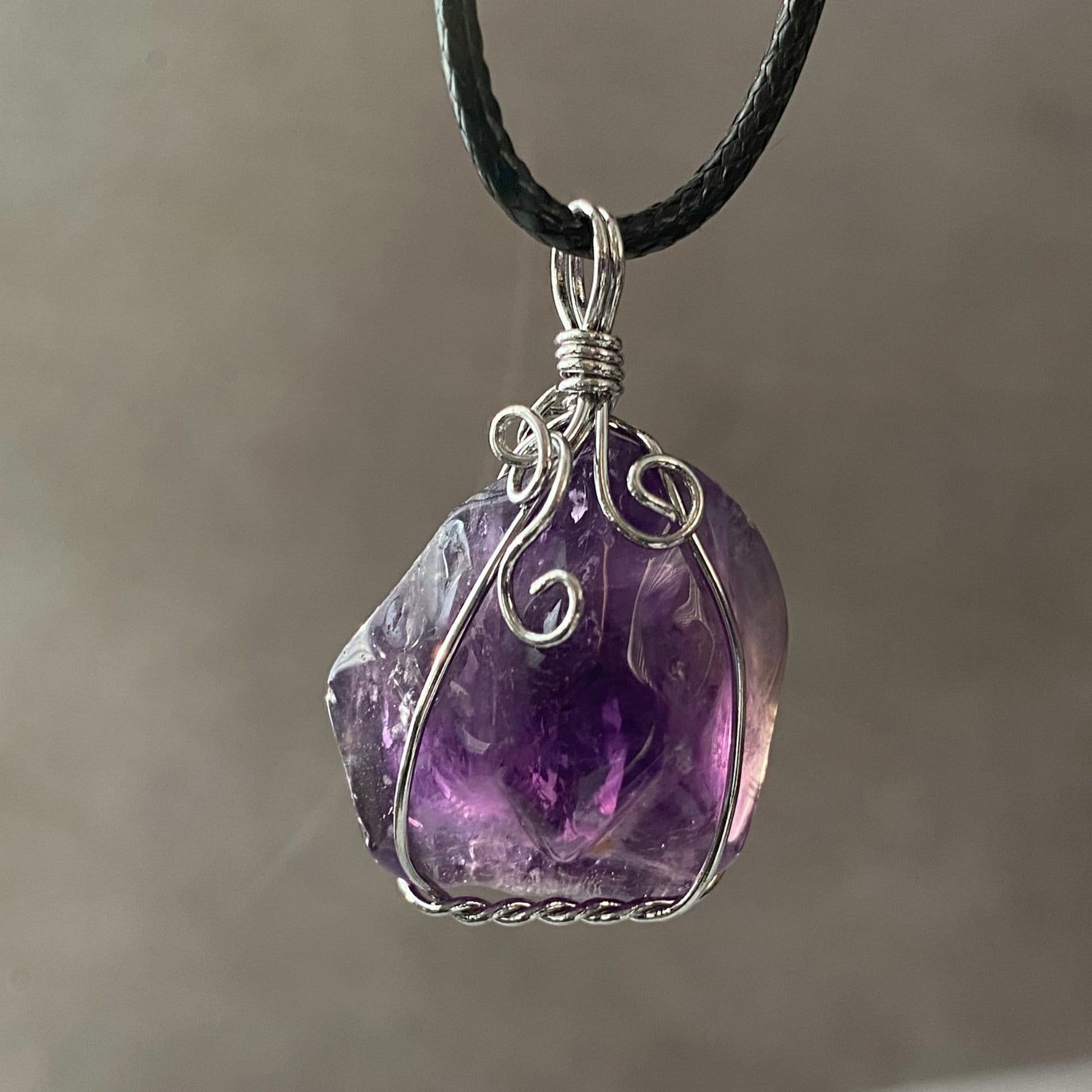 A close-up of a 1.25" Wire Wrapped Rough Amethyst Crystal Pendant wrapped in intricate silver wirework, hanging on a black cord. The crystal displays varying shades of purple. The blurred, neutral-toned background highlights the pendant's details. This piece not only enhances spiritual awareness but also wards off negative energies.