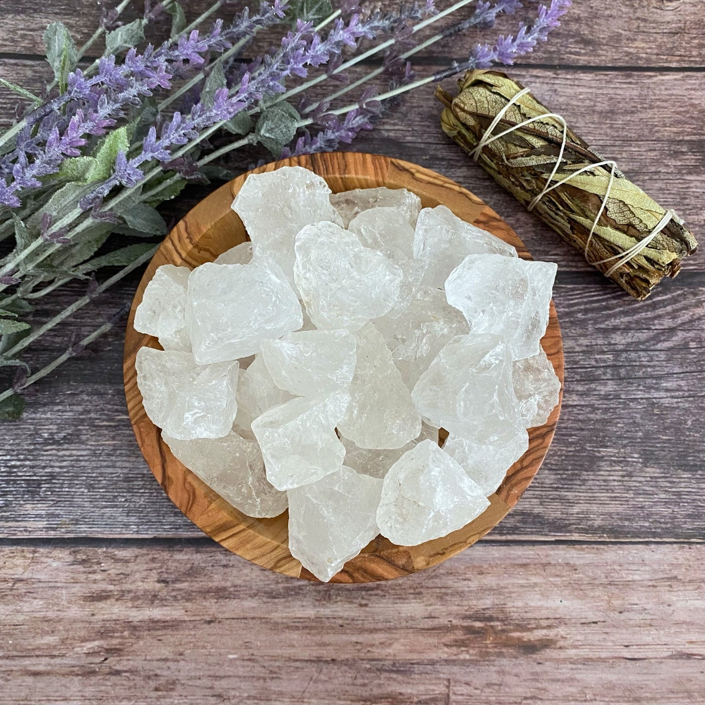 A wooden bowl filled with rough, white crystals is placed on a wooden surface. To the left, there are lavender sprigs, and to the right, a bundle of sage wrapped with twine. The Raw Clear Crystal Quartz Stones add to the arrangement's natural, rustic ambiance by amplifying energies for crystal healing.