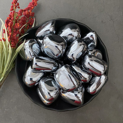 A black bowl filled with shiny, polished Terahertz Tumbled Stones sits on a dark, textured surface. To the left of the bowl, a small bundle of red berries and green foliage adds a touch of natural color. The arrangement not only captivates the eye but also serves as elegant EMF protection with its healing stones.